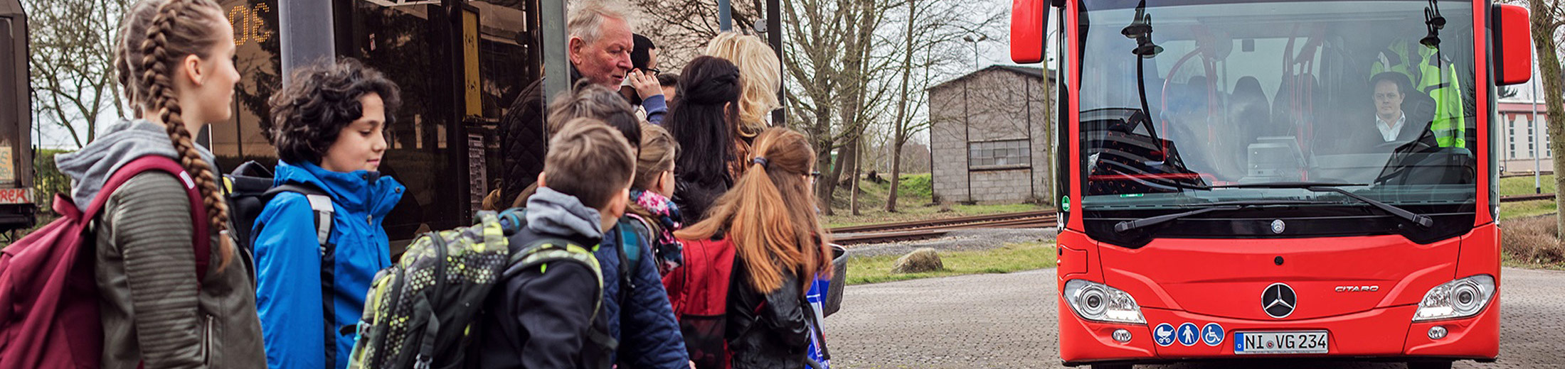 Dieses Bild zeigt mehrere Kinder und Erwachsene an einer Bushaltestelle. Im Hintergrund kommt ein Bus